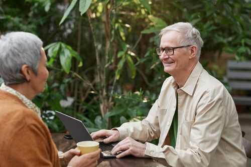 Two elderly individuals enjoying a conversation over coffee while using a laptop in a lush outdoor setting.