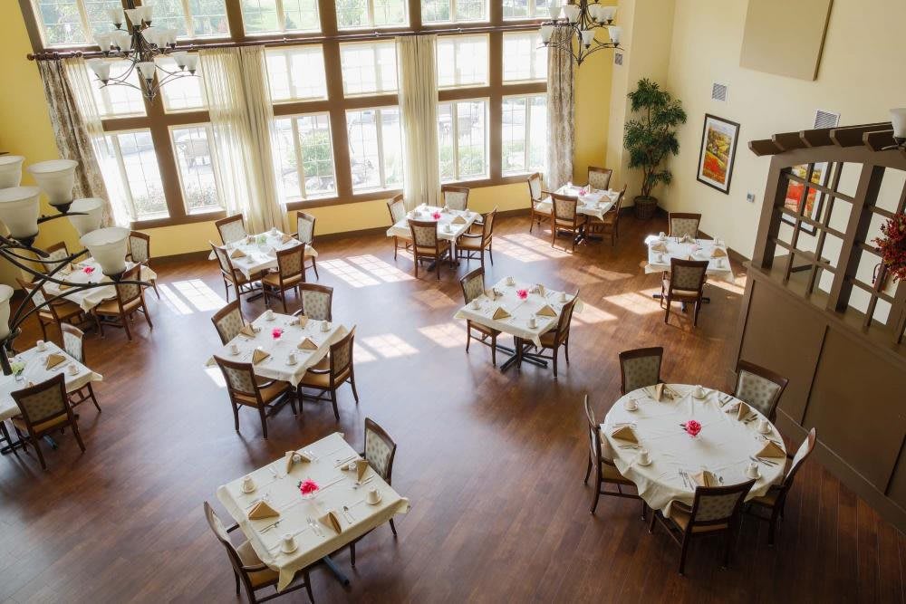 A sunlit dining area with neatly arranged tables and chairs, featuring large windows.