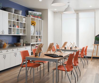 A brightly lit art classroom with orange chairs and easels set up on tables.