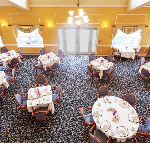 An elegant dining room with multiple tables set for a meal, featuring chandeliers and large windows.
