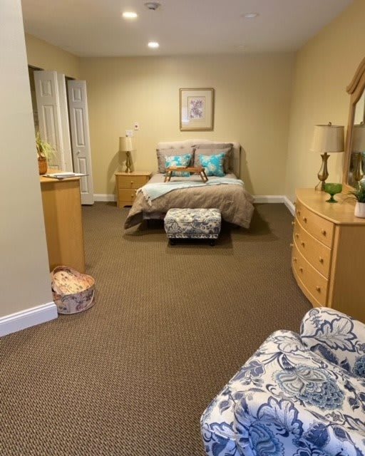 A cozy bedroom featuring a queen-sized bed with teal pillows, matching patterned ottoman and chair, and wooden furniture including dressers and tables.