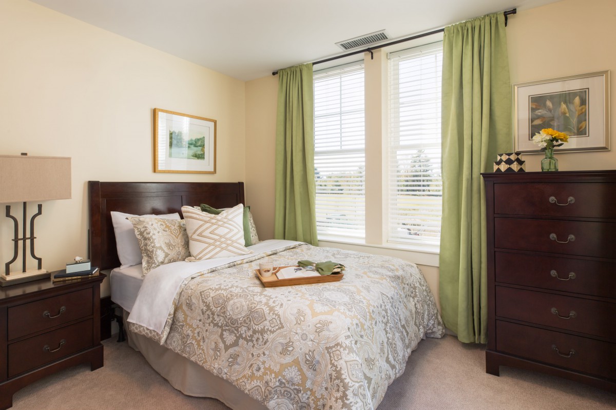 A cozy bedroom with a neatly made bed and green curtains letting in natural light through the windows.