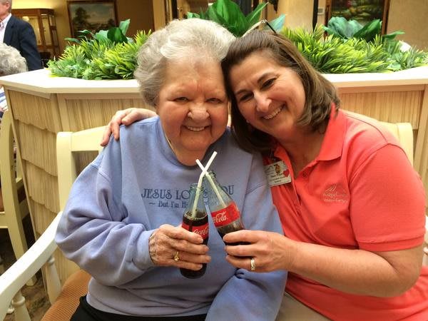 An elderly woman and her caregiver enjoy a moment together, sharing a smile and clinking Coke bottles.