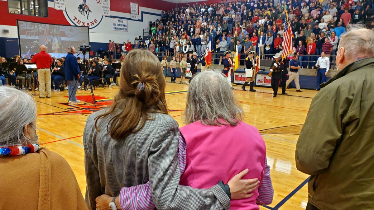 Two women embrace as a crowd gathers for a ceremony with flags in an auditorium.