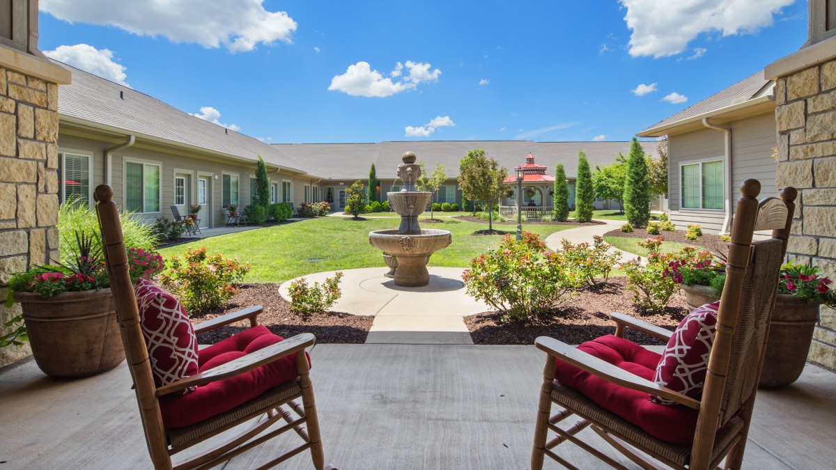 A sunny courtyard featuring a central fountain, surrounded by a neatly manicured garden and two red-cushioned chairs.