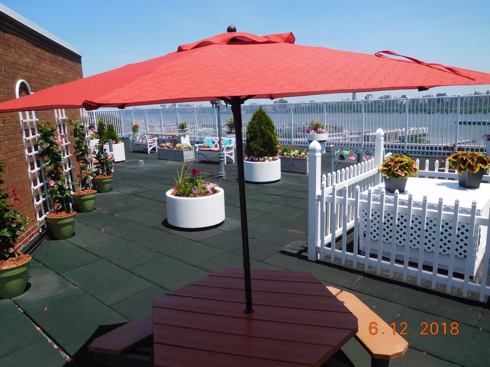 A rooftop patio with a red umbrella, surrounded by potted plants and a white fence overlooking the city.