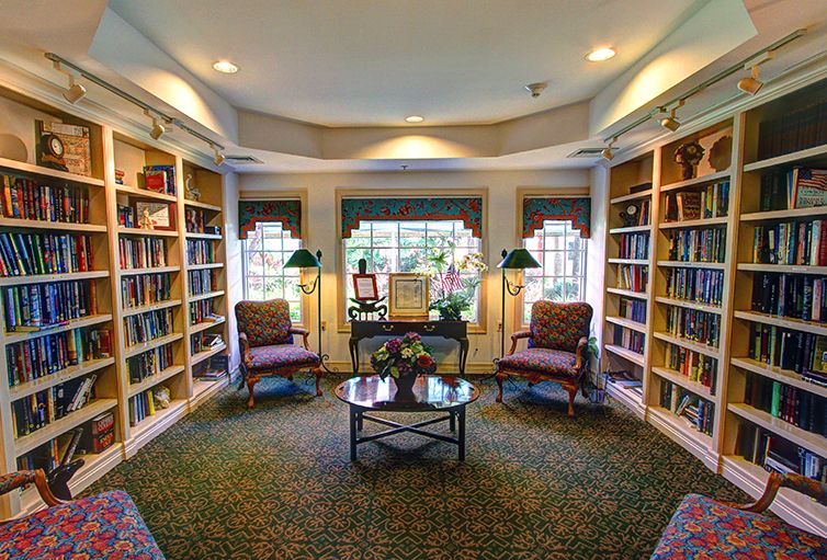A cozy library room with bookshelves, two chairs, and a central table beneath large windows.
