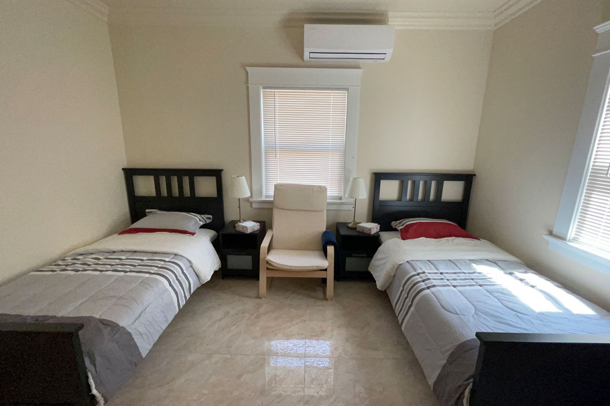 A minimalist bedroom features two twin beds, a chair, and an air conditioner above a window.