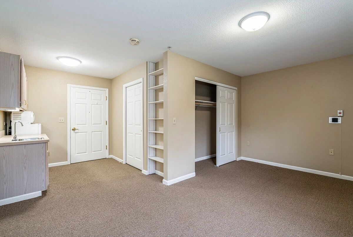 An empty room with beige walls, carpet flooring, and an open closet with built-in shelves.