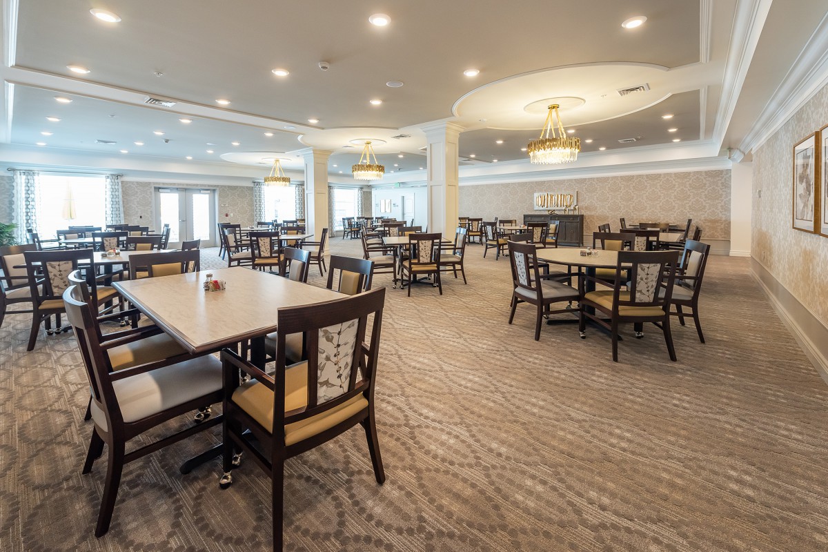 A spacious and well-lit dining hall with arranged tables and chairs, featuring chandeliers and a carpeted floor.
