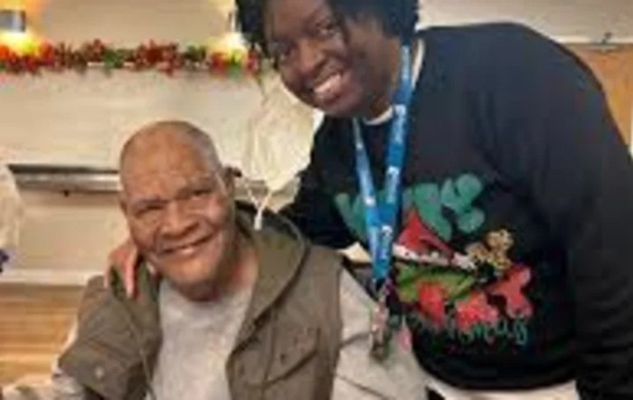 A smiling elderly man is seated next to a woman who is standing and leaning in for a photo.