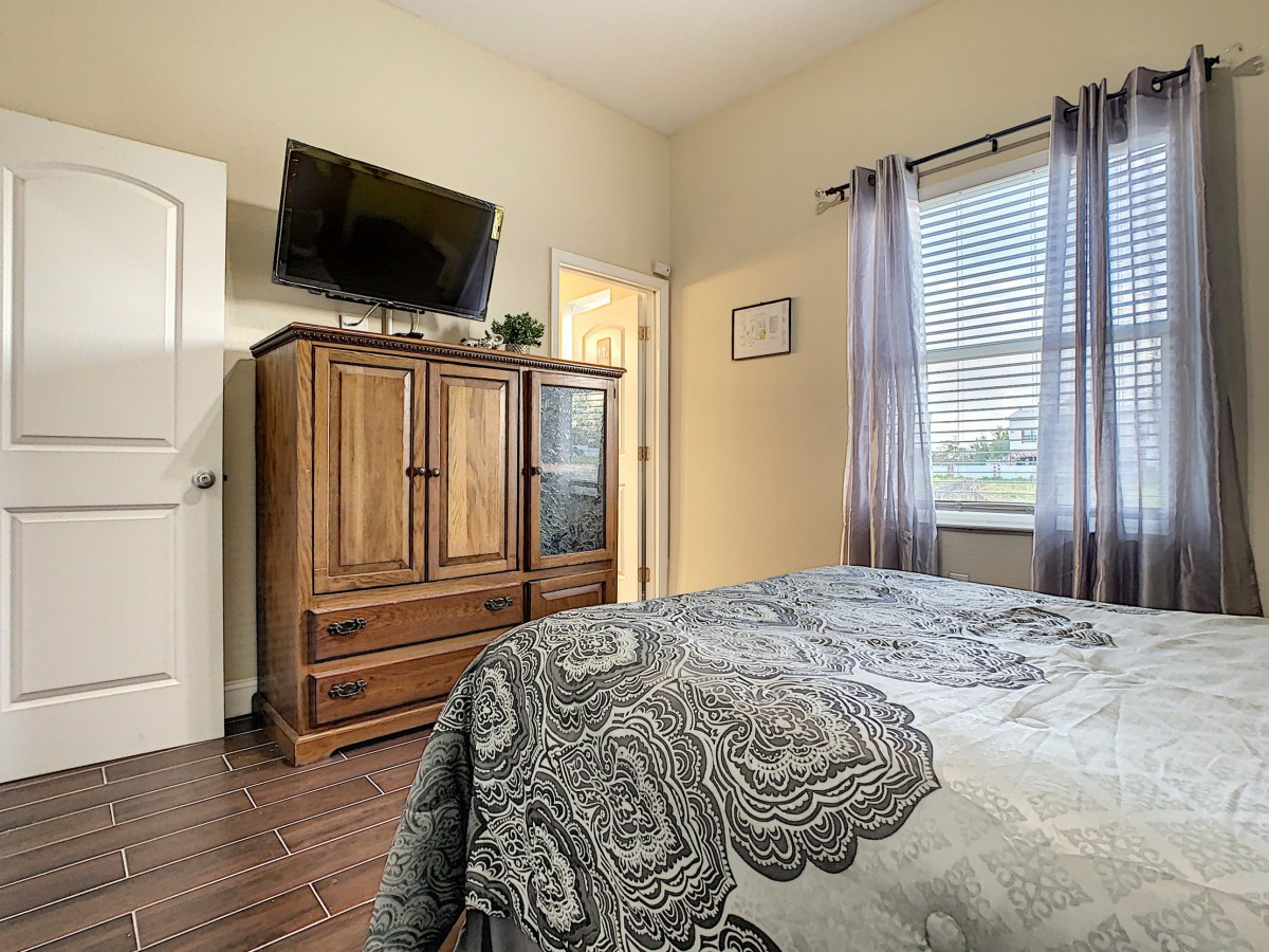 A cozy bedroom with a television mounted above a wooden armoire, featuring a bed with patterned bedding and a window with curtains.