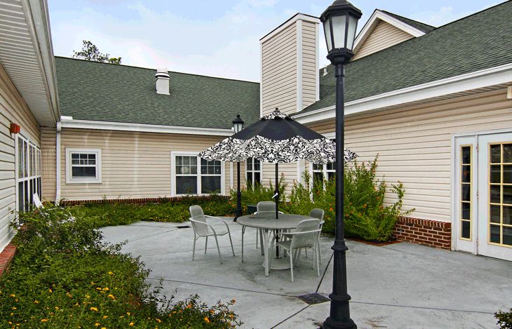 A quaint outdoor patio area with a table and chairs beneath an umbrella, surrounded by greenery and a lamp post.