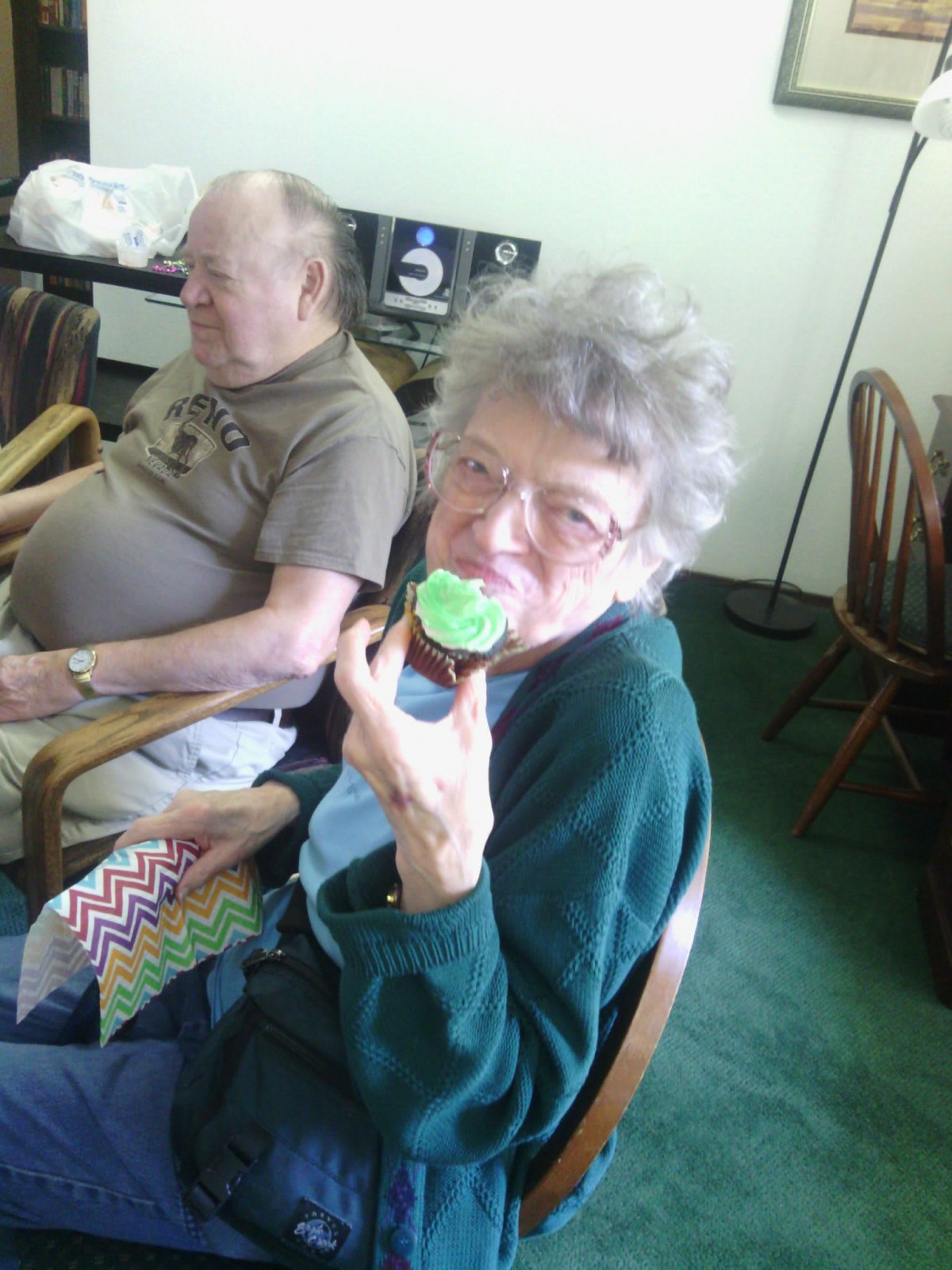 An elderly woman enjoying a cupcake while seated next to an elderly man indoors.