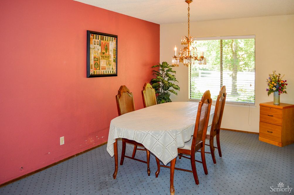 A cozy dining room with a table and four chairs, featuring a chandelier, artwork on a pink wall, and a window allowing natural light.