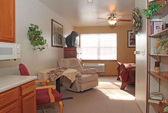 A cozy living room with an armchair, table, and ceiling fan, illuminated by natural light from the window.