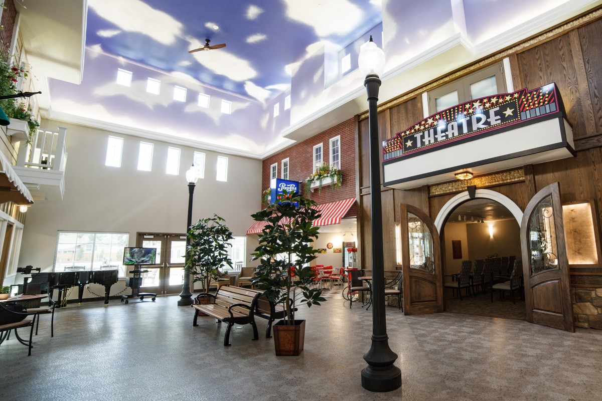 A spacious indoor atrium featuring faux storefronts, a cloud-painted ceiling, and decorative elements such as trees and lampposts.