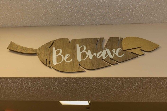 A wooden feather-shaped sign with the words 'Be Brave' displayed on a wall.