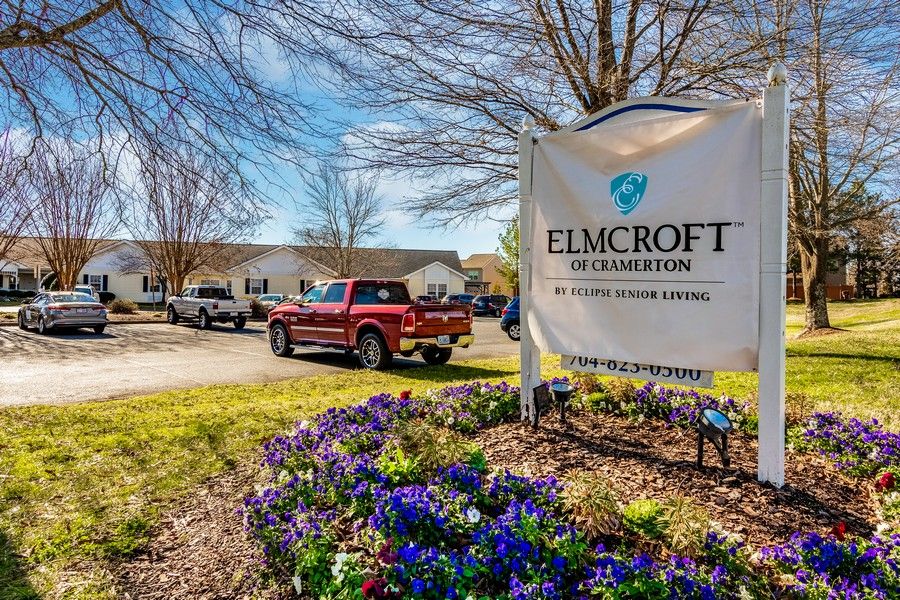 An entrance sign for Elmcroft at Cramerton with a parking area and blooming flowers.