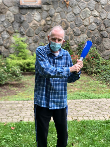 An elderly man wearing a mask stands outdoors, holding a blue object and posing in front of a stone wall.