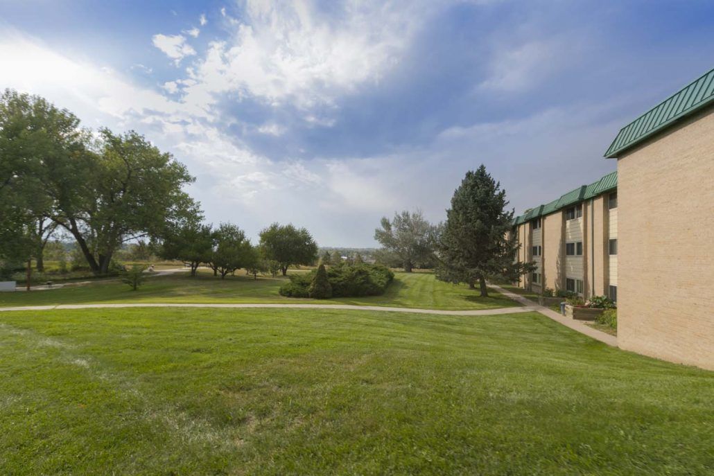 A scenic view of an apartment complex with a lush green lawn and trees under a partly cloudy sky.