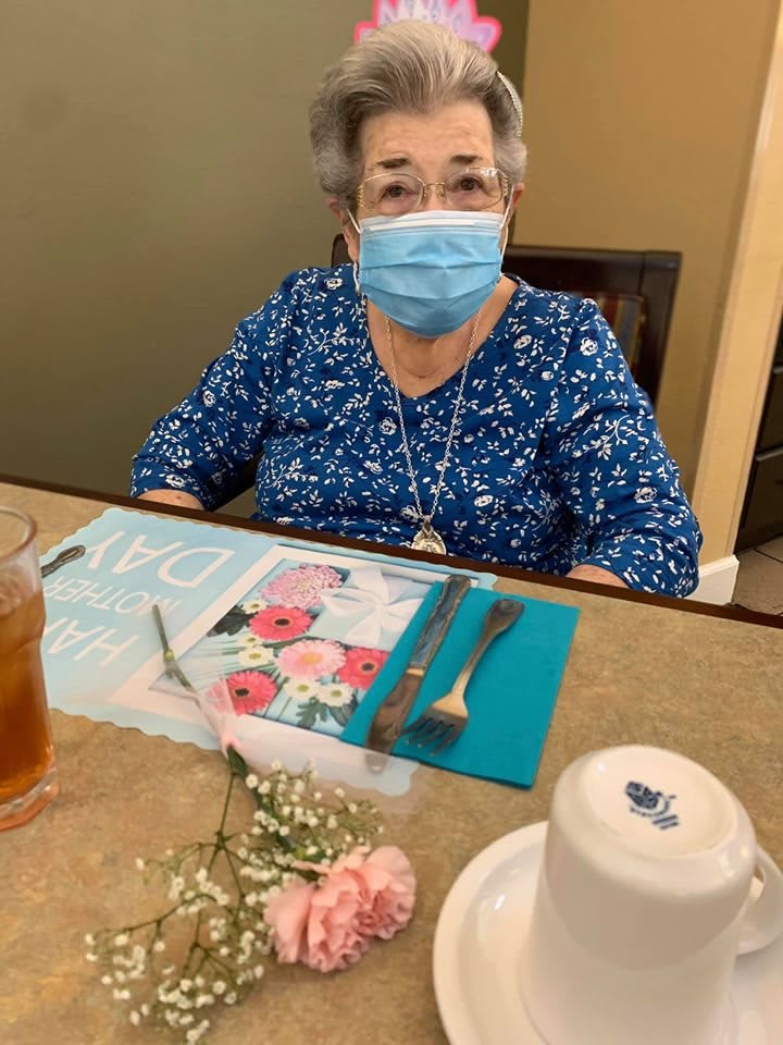 A woman in a blue shirt wearing a mask sits at a dining table with a flower and a drink.