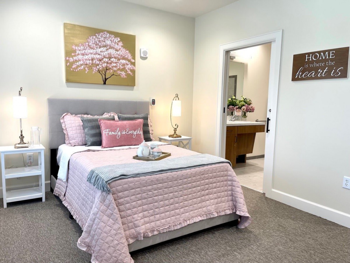 A cozy bedroom with pastel decor, featuring a bed adorned with pink and gray bedding, a side table with a lamp, and artwork of a cherry blossom tree on the wall.