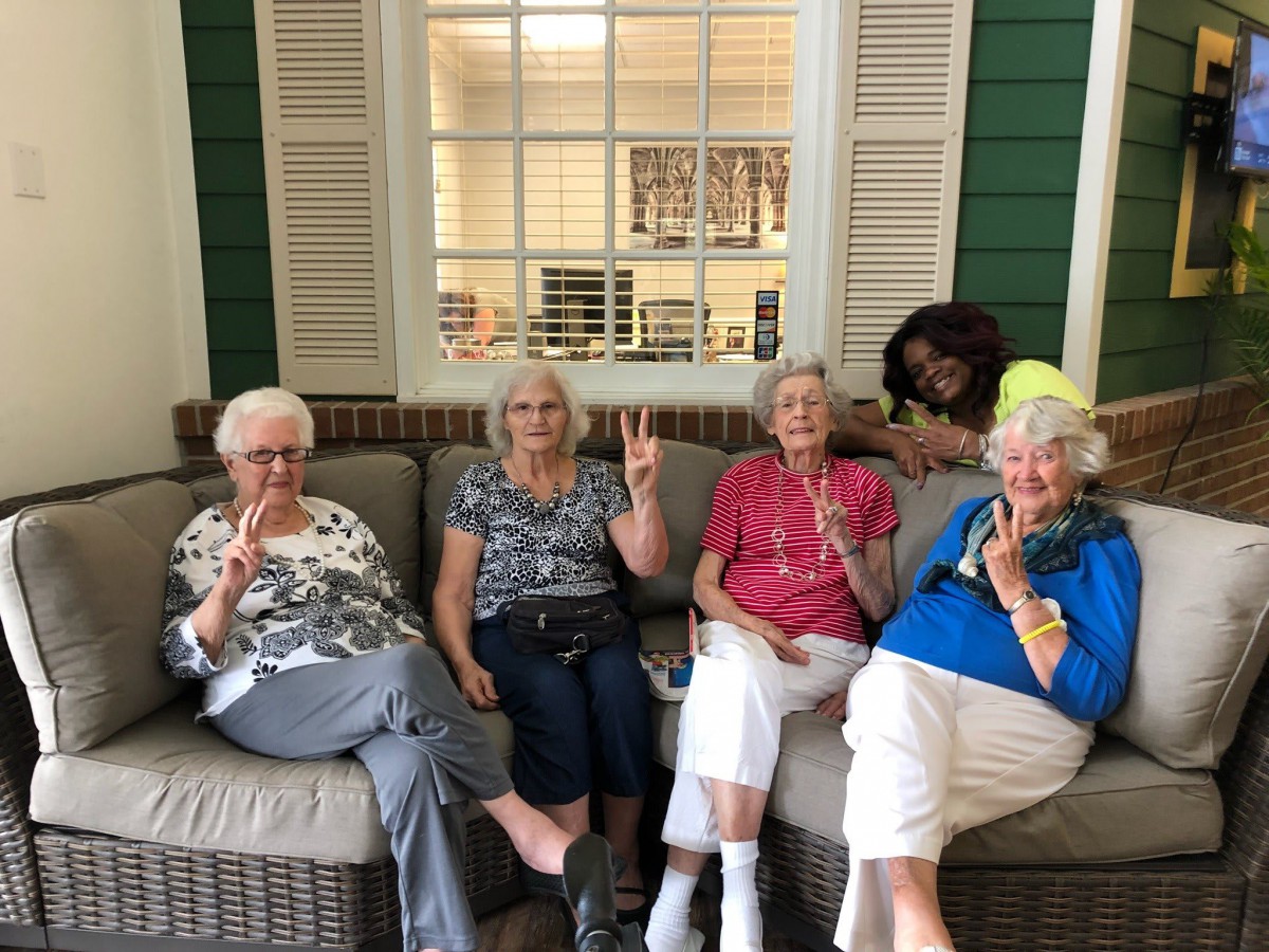A group of senior women sitting on a sofa indoors, showing peace signs.