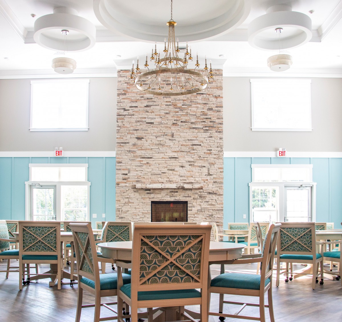 A spacious and well-lit dining area with a central stone fireplace, elegant chandelier, and neatly arranged tables and chairs.