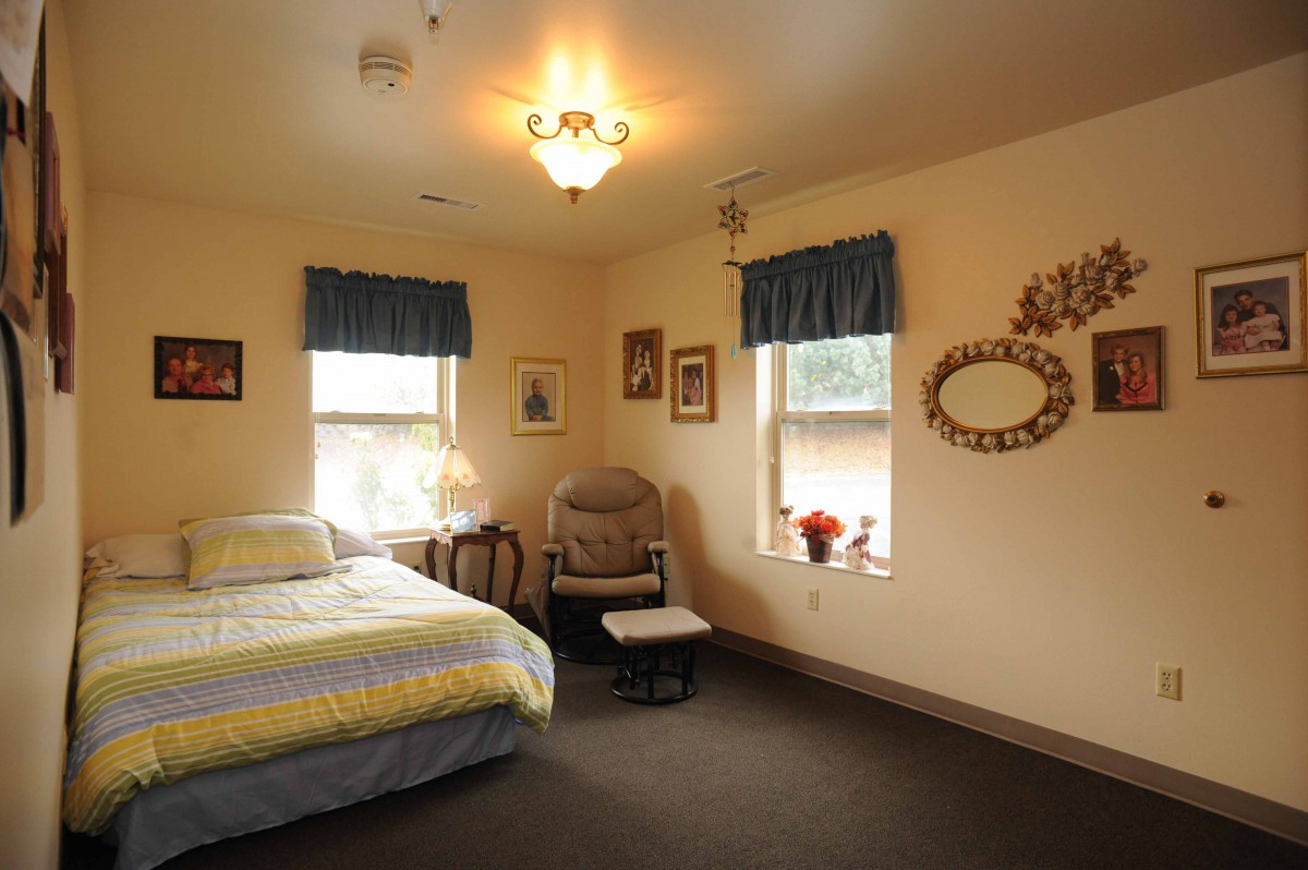 A cozy bedroom with a bed, chair, and wall decorations, softly lit by a ceiling light.