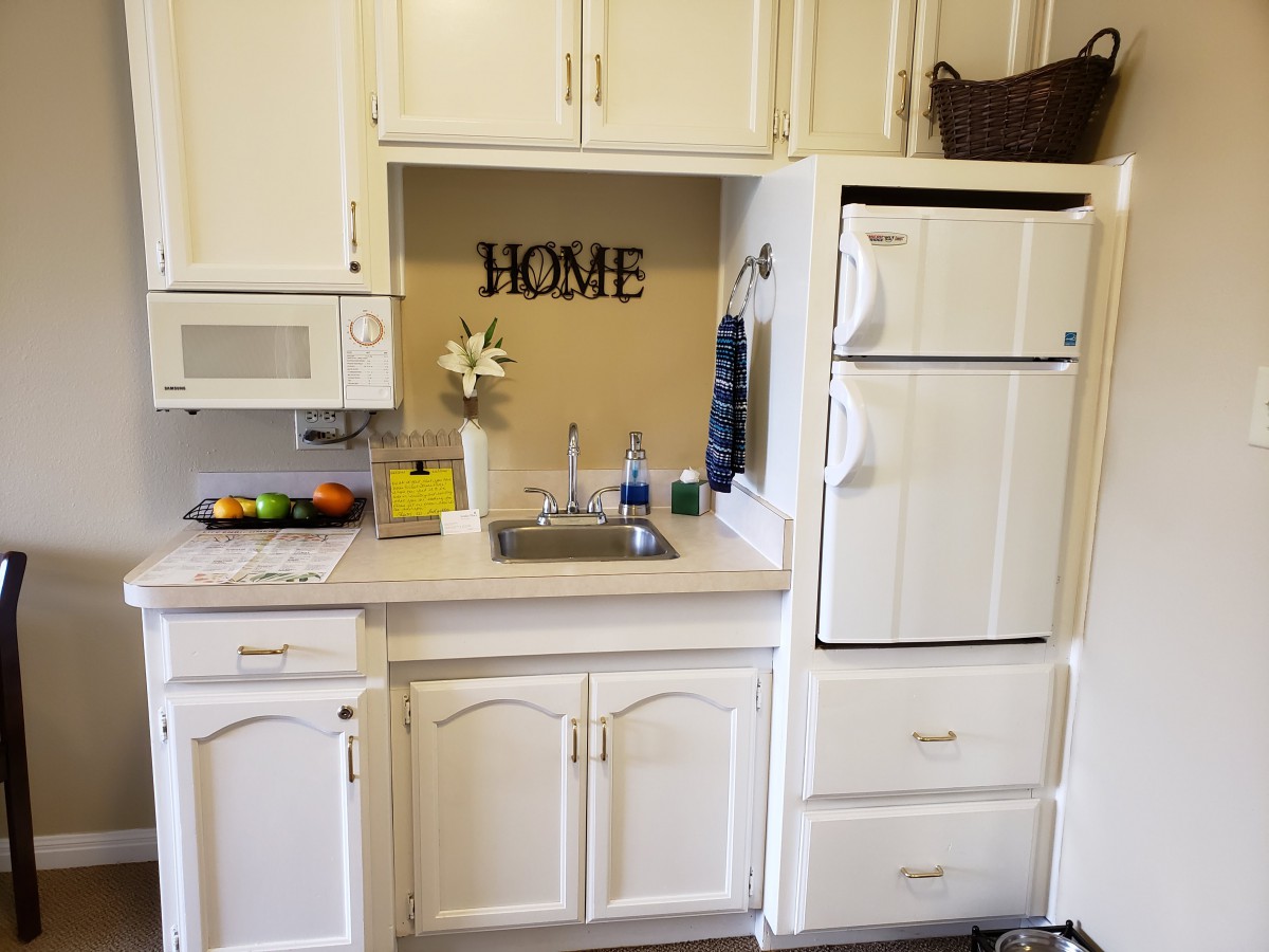 A cozy kitchen area featuring white cabinets, a sink, a refrigerator, and a microwave, with a decorative 'HOME' sign on the wall.