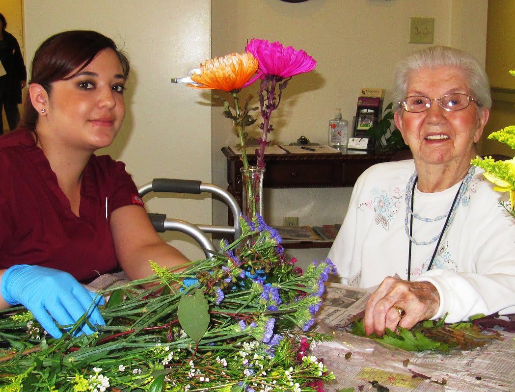 A young woman assists an elderly woman with arranging flowers at a table indoors.
