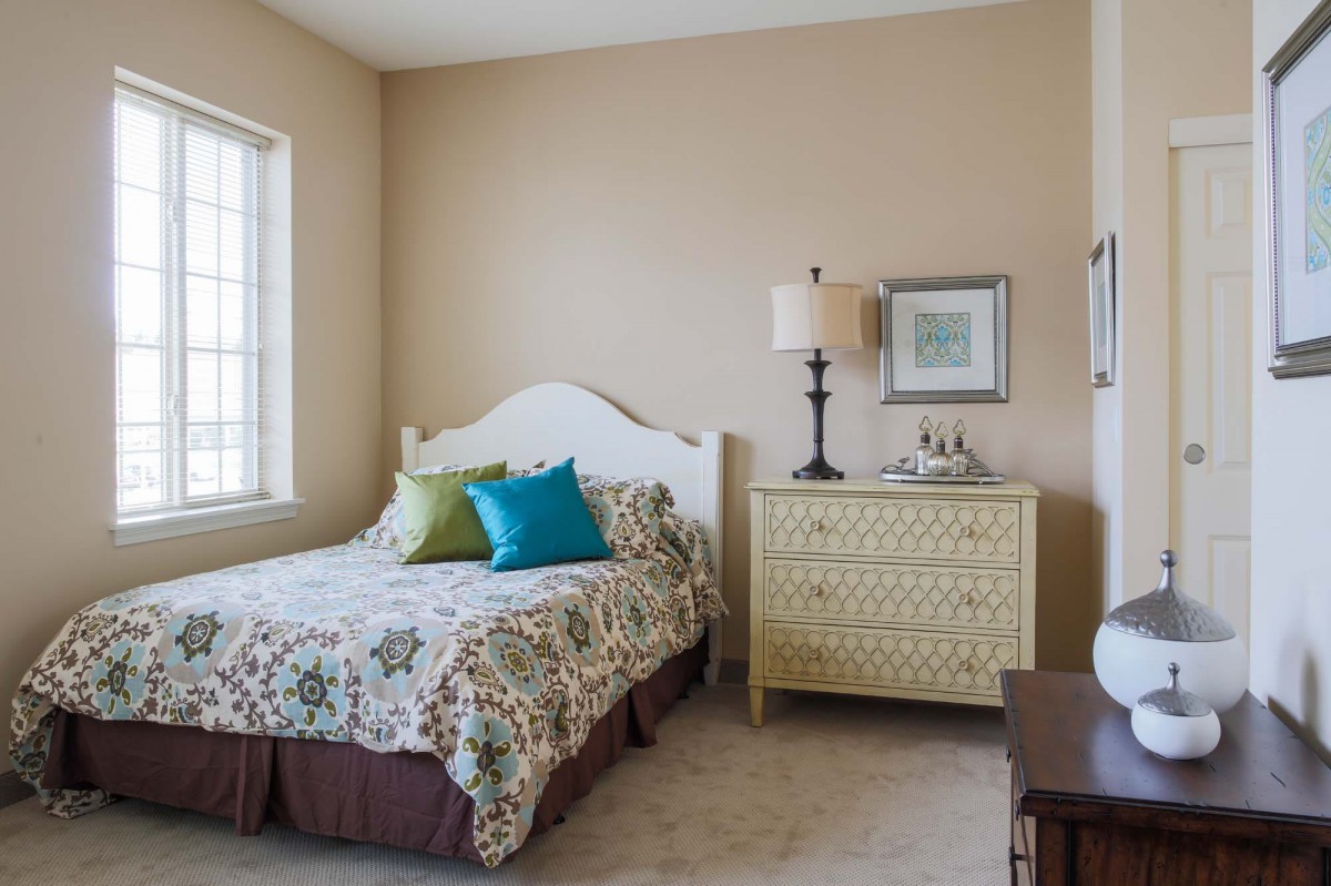 A cozy bedroom with a bed, colorful pillows, and a decorative dresser beside a window.
