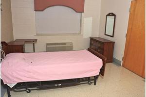A simple room with a bed covered in a pink sheet and basic furniture.