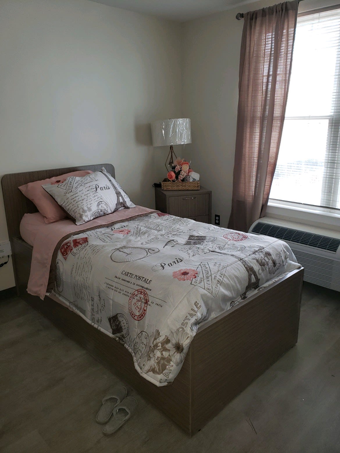 A neatly arranged bedroom featuring a single bed with floral bedding, a bedside lamp, and a window with sheer curtains.