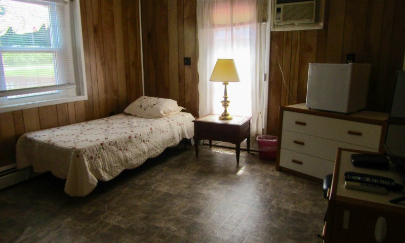 A cozy bedroom with a single bed, lamp, and dresser beside a curtained window.