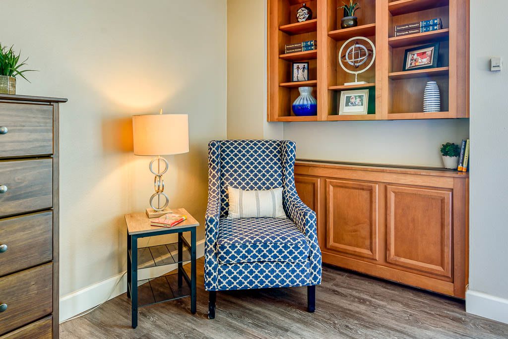 A cozy living room corner with a patterned armchair, side table, lamp, and built-in bookshelf.