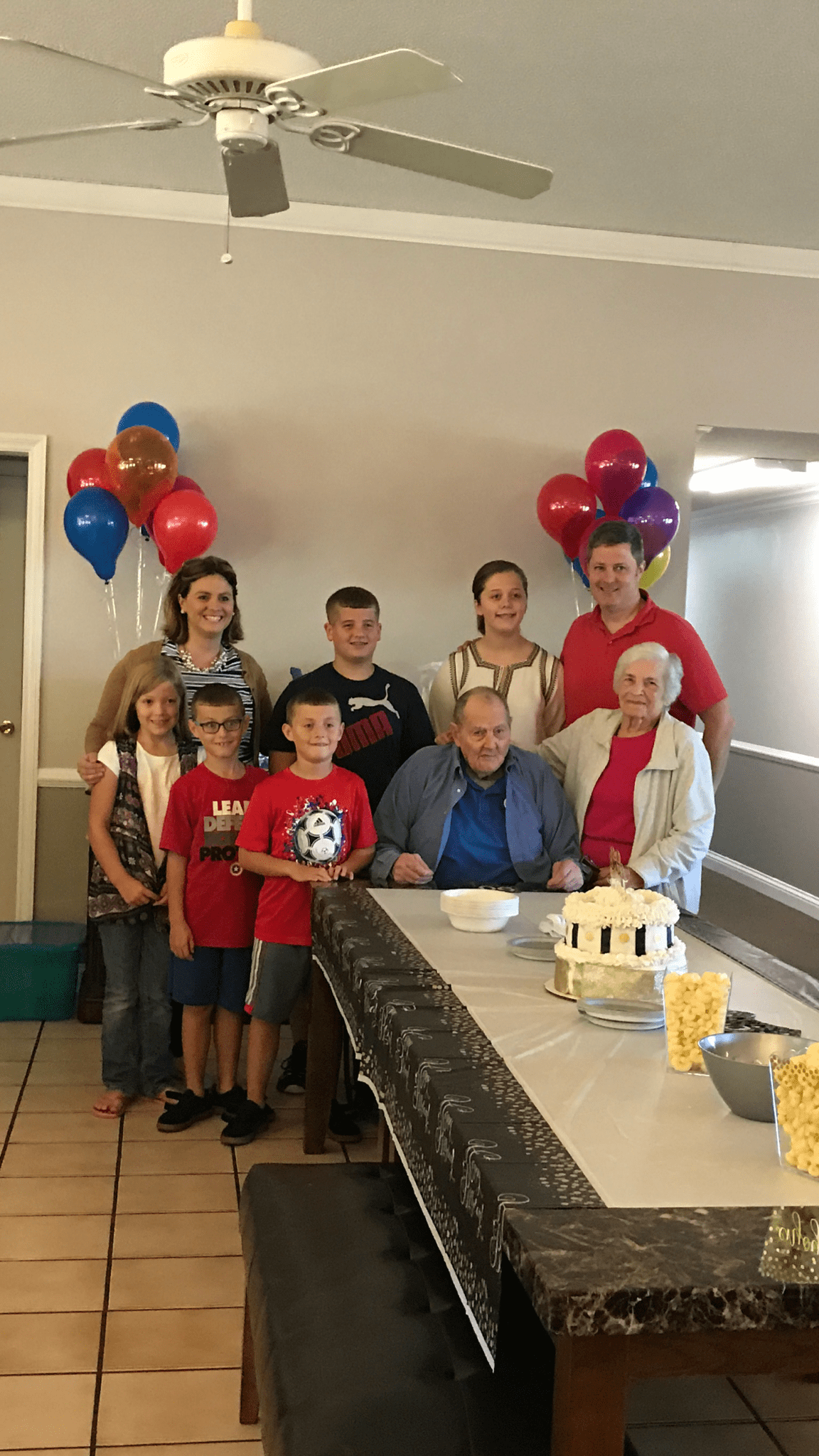 A family gathering around a table with balloons and a cake, celebrating a birthday.