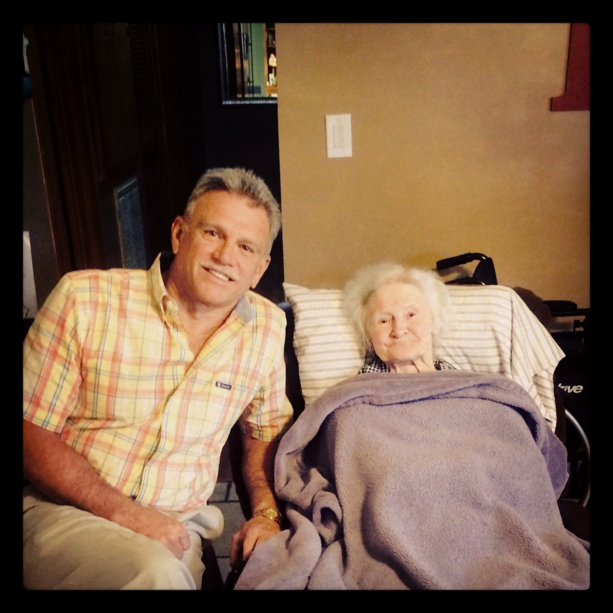 A person sitting next to an elderly woman covered with a blanket, indoors.