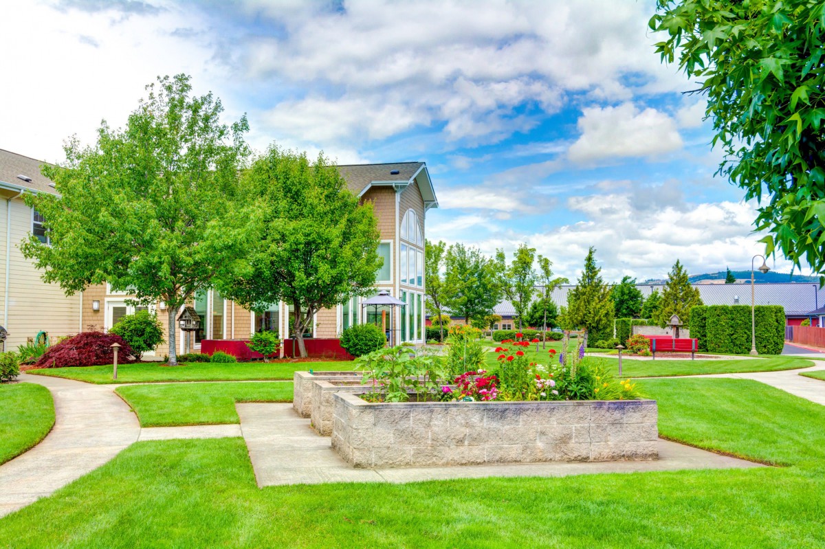 A scenic view of a garden with blossoming flowers, lush green grass, and a building in the background under a vibrant blue sky.