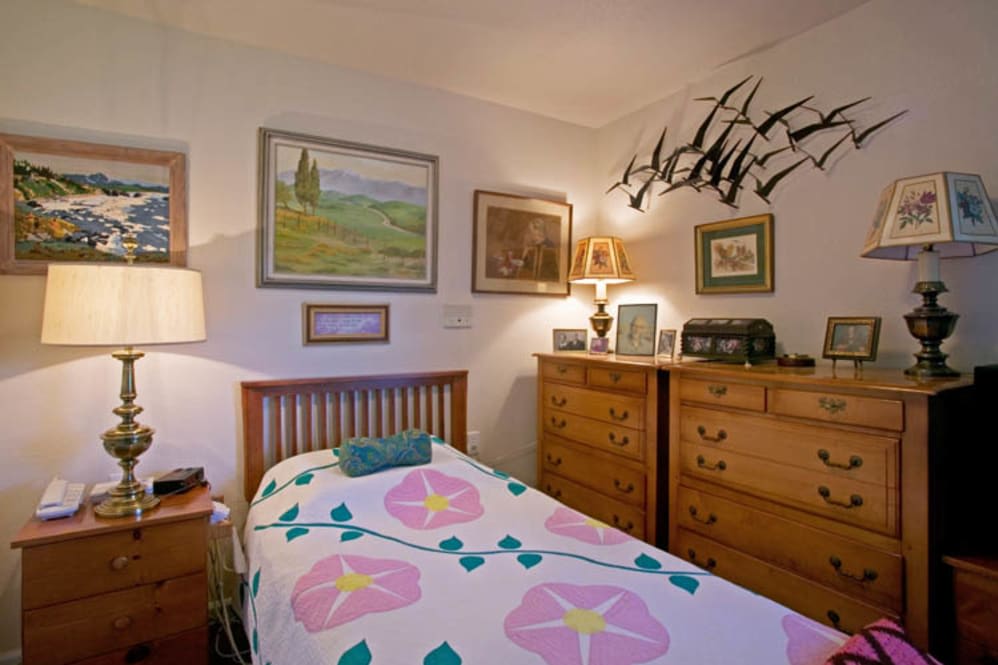 A cozy bedroom with a single bed adorned with a floral quilt, surrounded by artwork and wooden furniture.