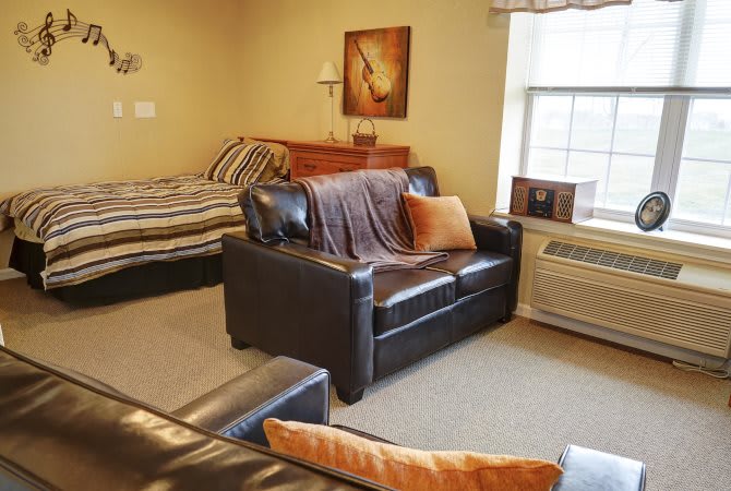 A cozy studio apartment with a combined living and bedroom space, featuring a leather sofa, bed, window, and air conditioner.