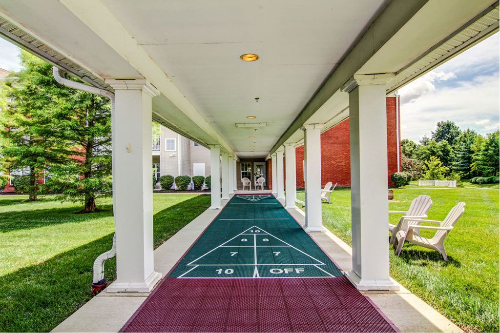 A covered outdoor walkway features a shuffleboard court with green and burgundy tiles, flanked by columns and surrounded by greenery.