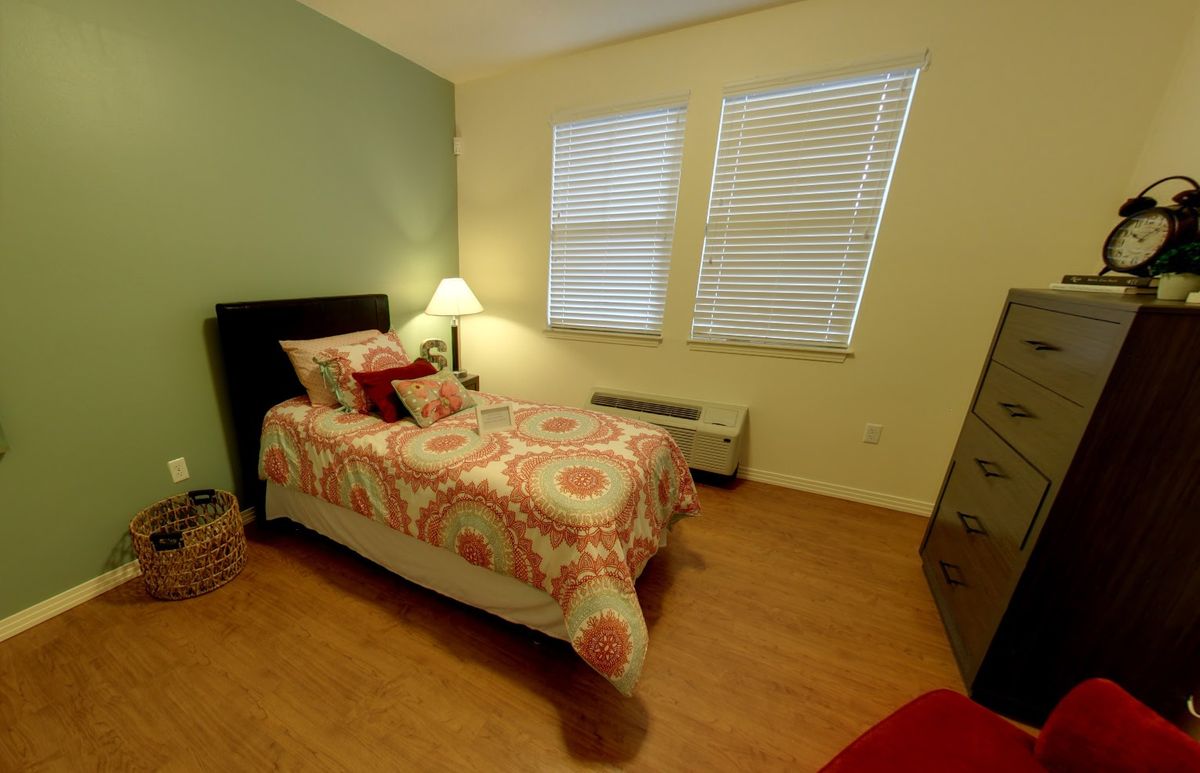 A cozy bedroom with a single bed, decorated with patterned bedding and cushions. The room features wooden flooring, a bedside lamp, and window blinds.