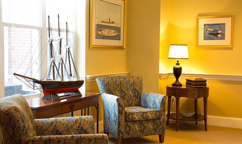 A cozy living room corner featuring an armchair, lamp, model ship, and artwork.