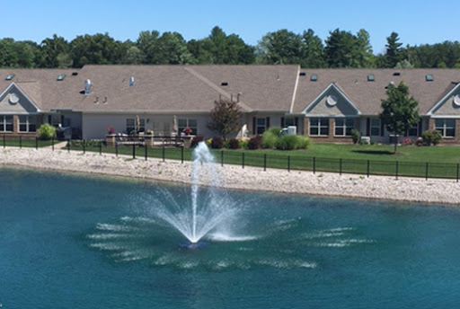 A scenic view of a water fountain in a pond, surrounded by a residential building and lush greenery.