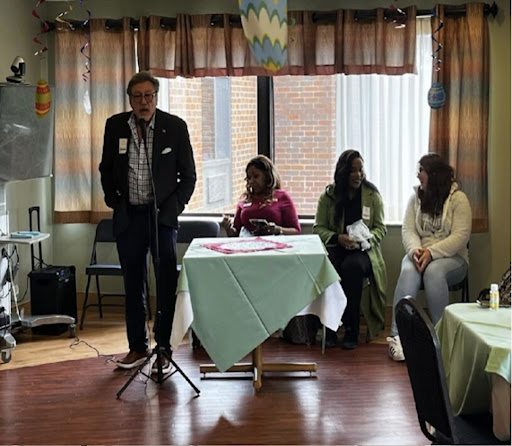 A speaker addresses the audience during a community event, with panelists seated nearby.
