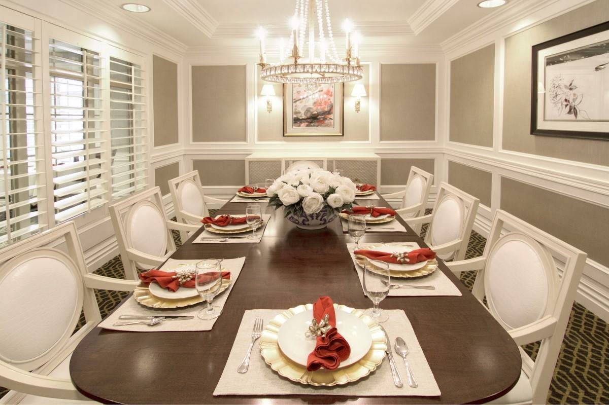 An elegant dining room with a beautifully set table featuring white and gold decor, red napkins, and a centerpiece of white flowers under a chandelier.