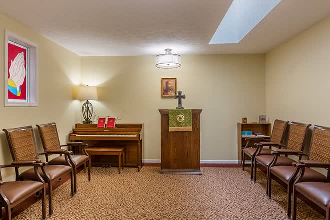 A small chapel room featuring a piano, chairs, and a pulpit with a cross.