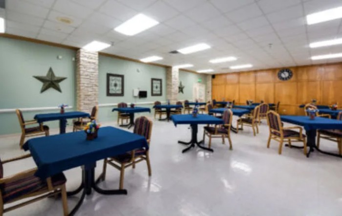 A spacious dining or meeting area featuring tables covered with blue tablecloths and several chairs, arranged in a well-lit room.